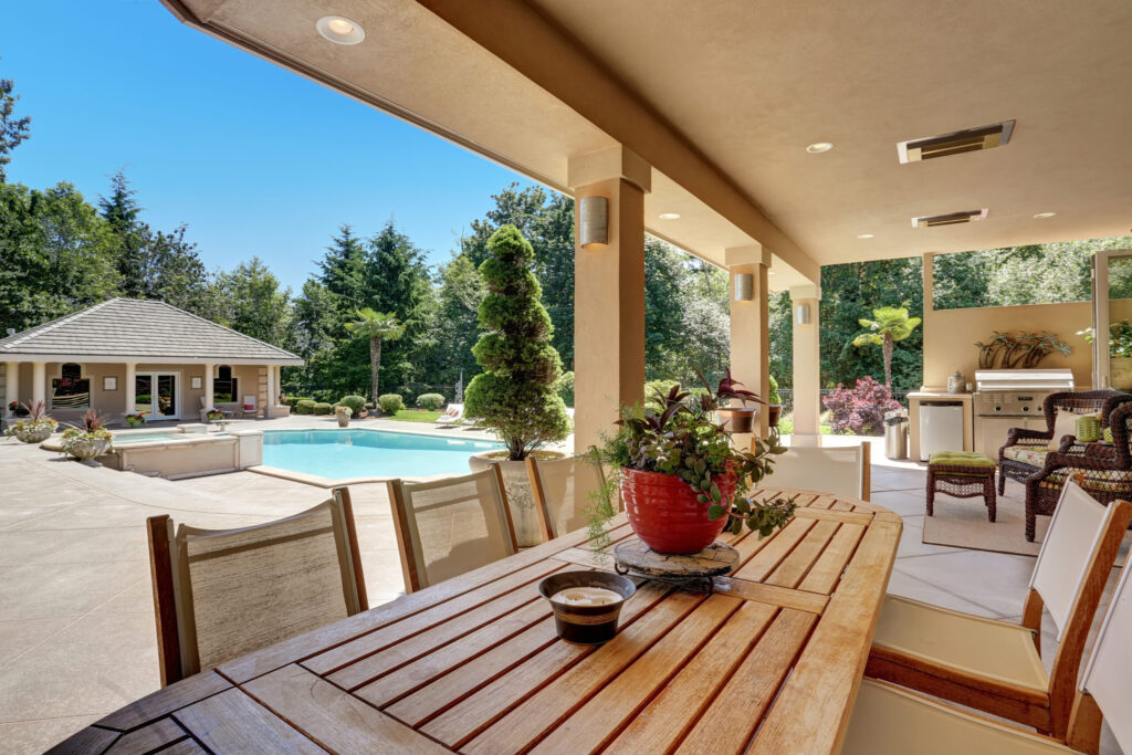 esterni di una casa in legno. si vedono una piscina, un gazebo, un tavolo in legno con 4 sedie e un vaso di margherite come centrotavola. il clima è soleggiato e fa pensare alla stagione estiva.